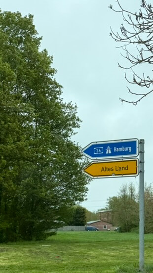 Wegweiser mit den Schildern „Altes Land" (gelb) und „A26 Hamburg" (blau) vor grünen Bäumen