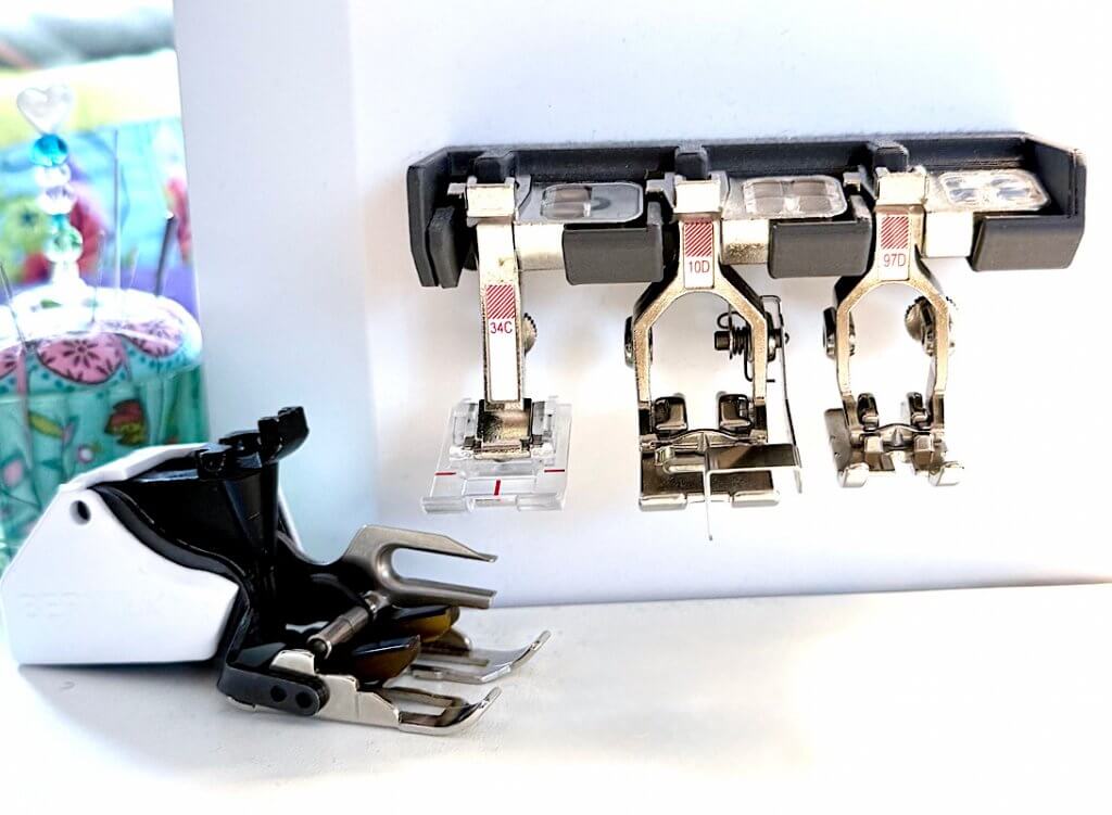BERNINA Nähmaschine mit Nähfußhalter, bestückt mit Fuß 34C (Klarsichtfuß), 10D (Schmalkantfuß) und 97D (Patchworkfuß); davor liegt der Obertransportfuß