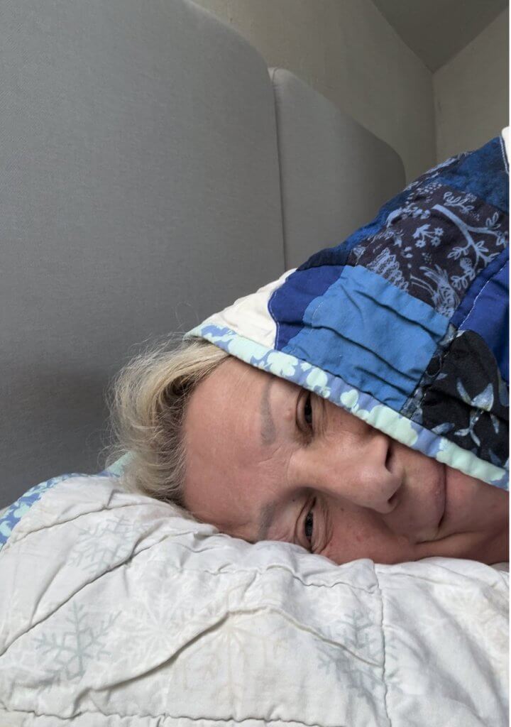 Person liegt im Bett, ein blauer Patchwork-Quilt ist bis über den Kopf gezogen