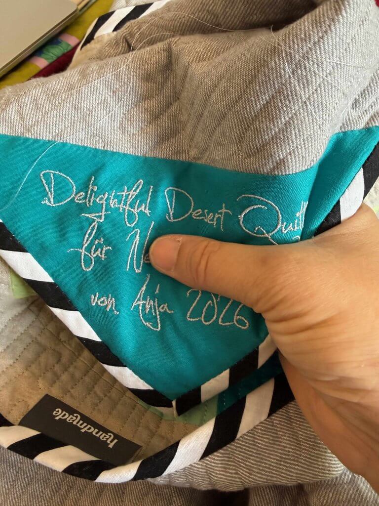 Türkisfarbenes, besticktes Quiltlabel mit Quiltnamen und Jahr, daneben ein Handmade-Weblabel auf der Quiltrückseite