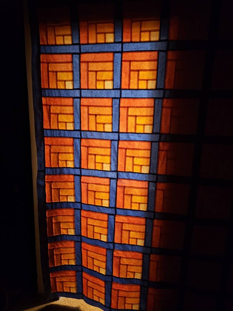 Lichtspiel-Quilt in Orange und Blau gegen das Licht fotografiert, die Blöcke leuchten wie ein Kirchenfenster