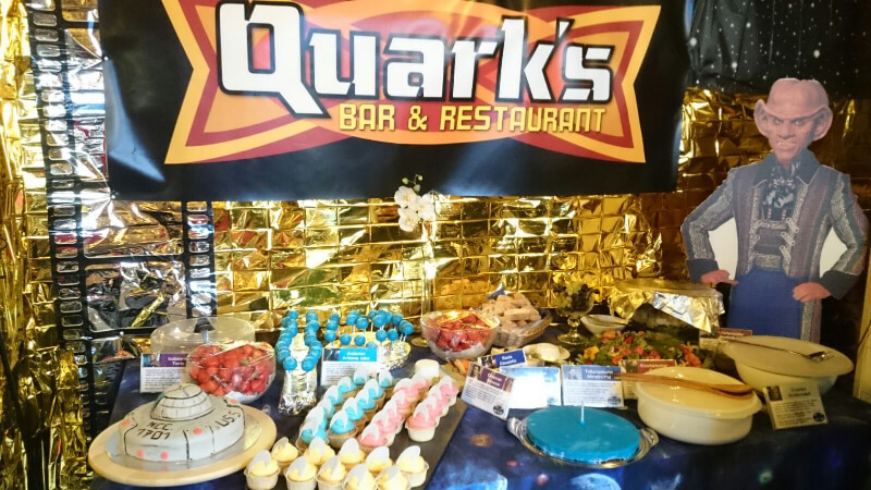 Buffet unter einem Banner „Quark’s Bar & Restaurant” mit Kuchen in Raumschiff-Form, blauen und rosa Törtchen, vor Wänden aus goldenen Rettungsdecken