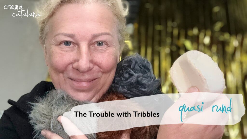 Anja Dix hält drei genähte Fellbälle aus Kunstfell in Hellgrau, Dunkelgrau und Rotbraun und ein eckiges Probemodell aus Schaumstoff vor goldenem Hintergrund, dazu der Titel „The Trouble with Tribbles” und der Schriftzug „quasi rund”